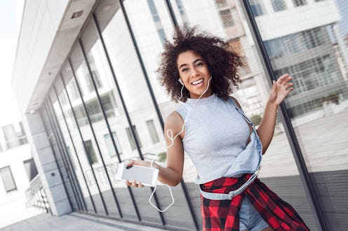 Young woman in the city street walking near window holding smartphone wearing earphones listening to...