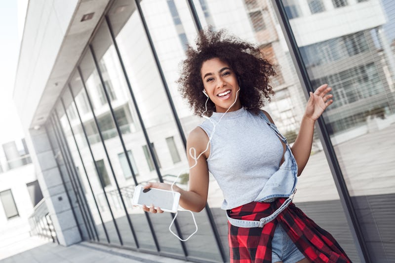 Young woman in the city street walking near window holding smartphone wearing earphones listening to...