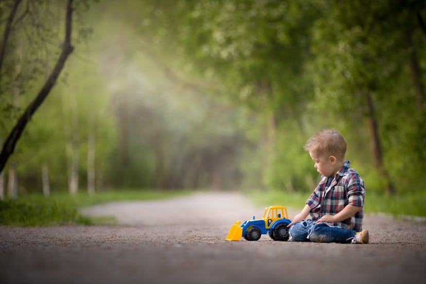 Portrait of cute kid boy sitting on the ground and playing with colourful tractor and sand in the pa...