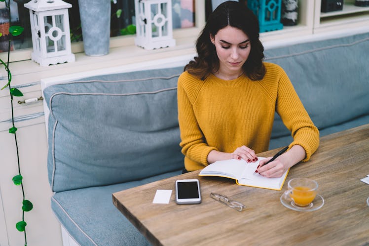 Young concentrated beautiful woman with brown hair in yellow sweater writing in notebook with empty ...