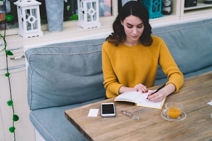 Young concentrated beautiful woman with brown hair in yellow sweater writing in notebook with empty ...