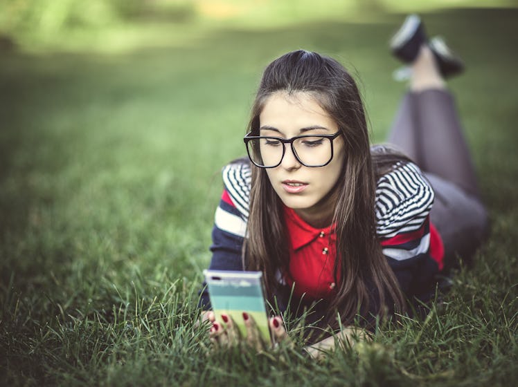 Beautiful young hipster girl using smart phone lying on the grass in the park