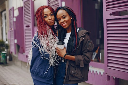 two beautiful and stylish dark-skinned girls with long hair standing in a autumn city near purple wa...