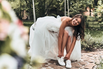 Perfect look! Attractive young woman in wedding dress putting on sports shoes and smiling while sitt...