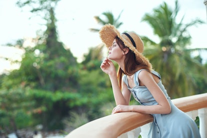 A dreamy woman in a sundress and a hat leaned on the balcony railing and looks at nature.        