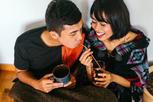 A young Malay couple snuggle up and share a hot beverage in their home. They are wrapped up and sitt...