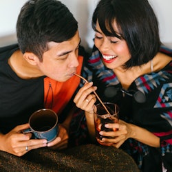 A young Malay couple snuggle up and share a hot beverage in their home. They are wrapped up and sitt...