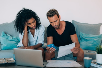 Young interracial couple in the couch stressed with financial problems doing calculations with paper...