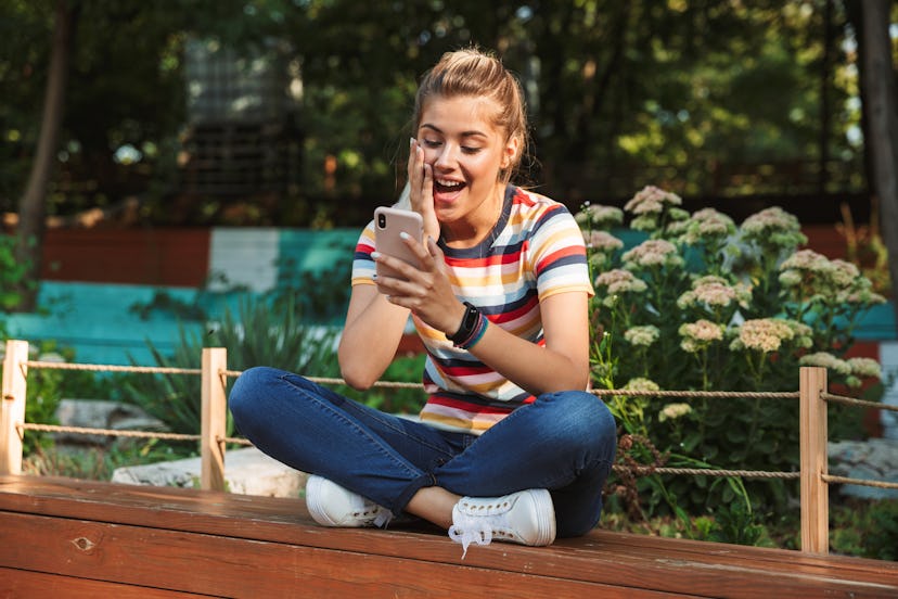 Portrait of a happy young teenage girl sitting on a bench at the park, looking a tmobile phone, laug...