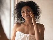 Happy african american young woman looking in mirror at bathroom, wrapped in towel after morning sho...