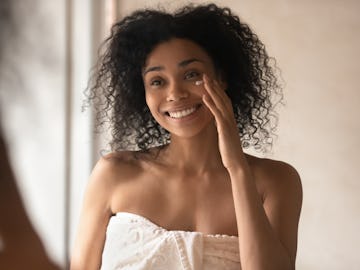 Happy african american young woman looking in mirror at bathroom, wrapped in towel after morning sho...