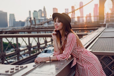 Beautiful traveling woman in red dress and stylish black hat enjoying amazing view from Brooklyn bri...