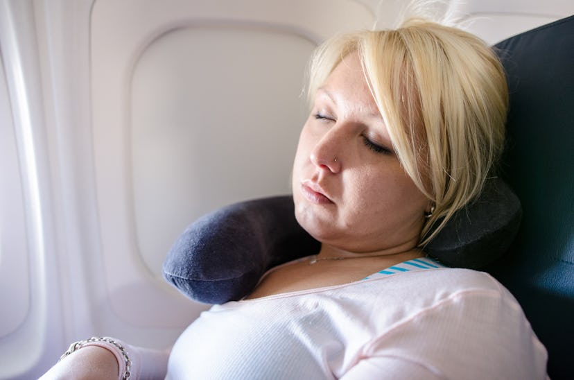 A blonde young adult woman uses a neck pillow to sleep and nap on a long airplane flight. Woman is i...