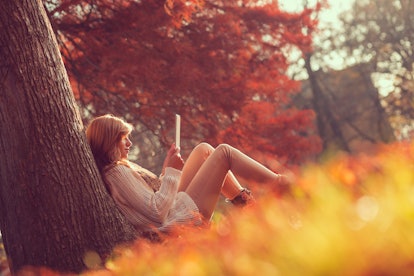 Beautiful young brunette sitting on a fallen autumn leaves in a park, reading an e-book on a tablet ...