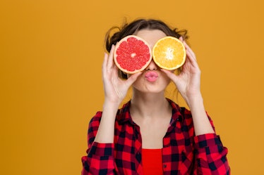 Funny playful young woman in checkered shirt holding halves of citrus fruits against her eyes and ma...