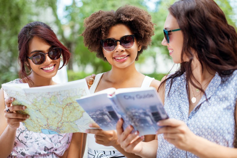 tourism, travel and friendship concept - happy tourist women or friends with map and city guide on s...