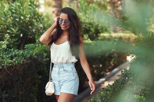 Young beautiful hipster casual dressed girl wearing shirt and jeans shorts walking between green pla...