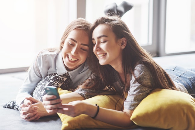 Two cute smiling twins sisters holding smartphone and making selfie. Girls lie on the couch posing and joy.
