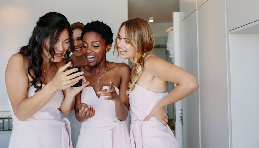 Smiling bridesmaids looking at the photographs of bride on mobile phone in a hotel room on wedding d...