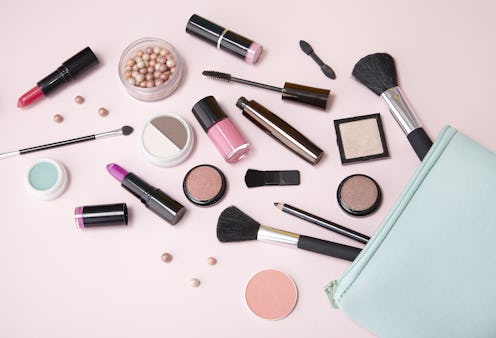 A blue make up bag with cosmetic beauty products spilling out on to a pastel pink background