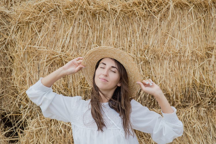 Young girl wears summer white dress near hay bale in field. Beautiful girl on farm land. Wheat yello...