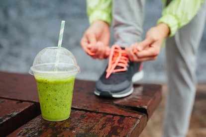 Green detox smoothie cup and woman lacing running shoes before workout on rainy day. Fitness and hea...