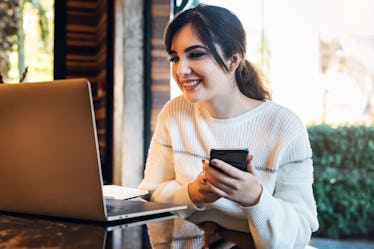 Portrait of smiling young woman with smartphone in her hands, sitting at table in front of laptop. G...