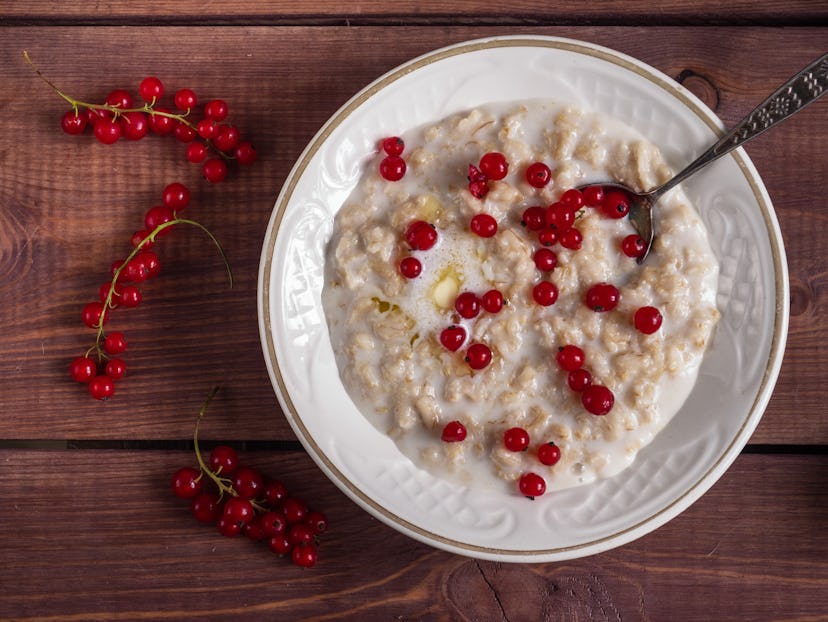 Oatmeal on milk with red currant in a white plate on a wooden brown tray, a scattering of currants o...