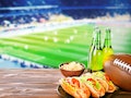 Beer with snack and ball on wooden table against football field background. Sport and entertainment ...