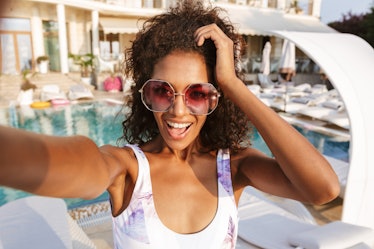 Cheerful young african woman in swimsuit taking a selfie while spending good time a swimming pool re...