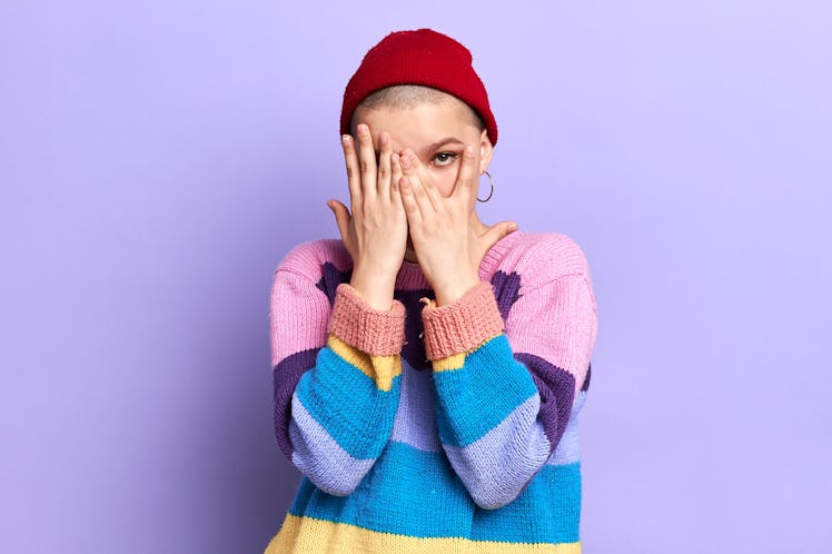 shy cool glamour girl in striped colorful sweater covers her face with her hands and looks through h...