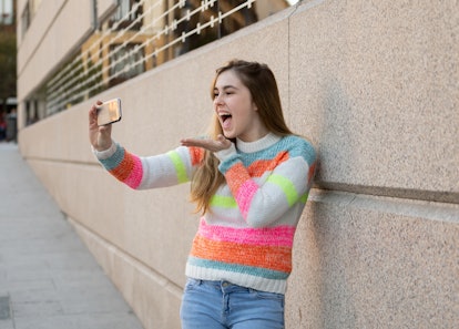 Happy pretty teenager girl using smart mobile phone excited blowing a kiss and waving to followers f...