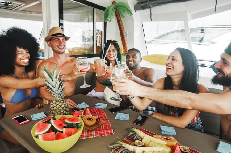 young friends drinking laughing saying cheers with glasses of cocktails and wine inside a boat.