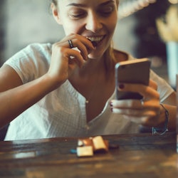 young pretty brunette woman eat chocolate in cafe, using smartphone, touch screen display, sweet foo...