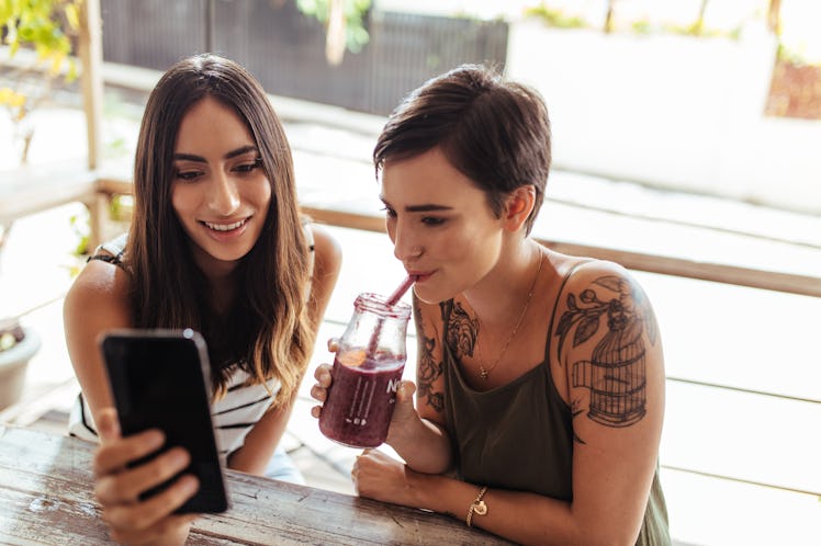 Two women sitting at a restaurant looking at a mobile phone. Woman showing mobile phone while anothe...