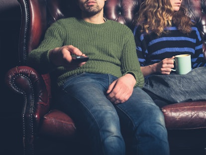 A bored young couple are sitting on a sofa at home watching television