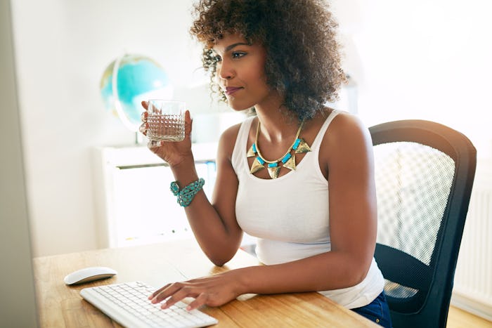 Young attractive African American businesswoman drinking water as she sits at her desk typing inform...