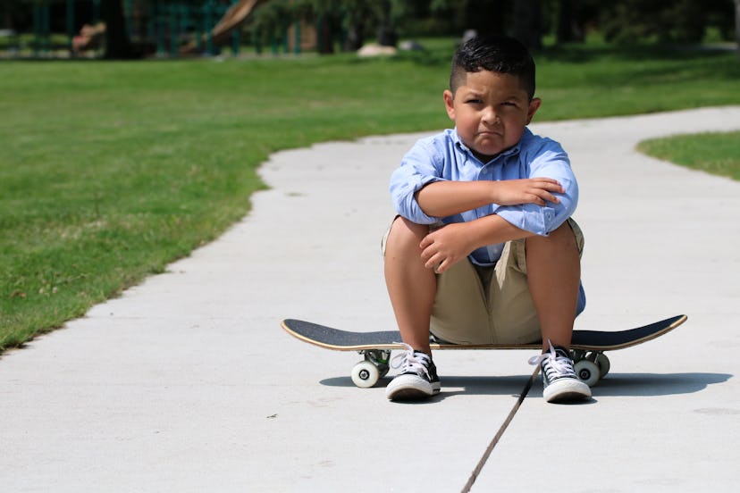 A young hispanic child is sitting on a skateboard in a park, upset or angry. 