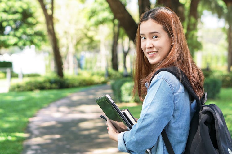 Portrait of asian female colleges student holding textbook in her hands and standing outdoors at uni...