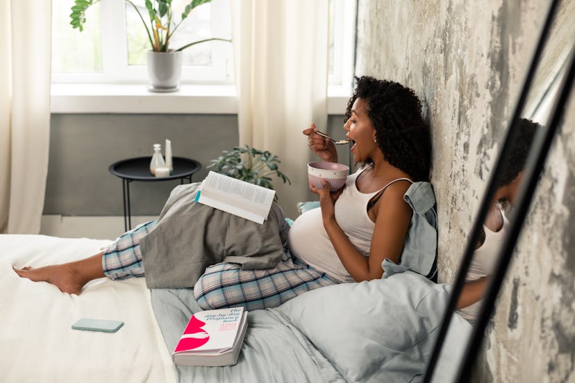 Information search. Smiling pregnant woman eating in her bed surrounding herself with books.