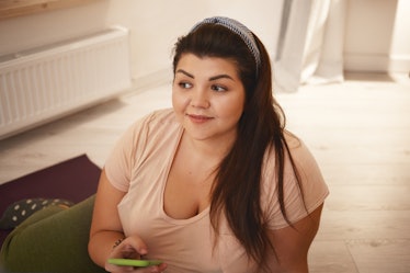 Close up shot of positive young obese female with chubby cheeks and curvy body typing text message v...