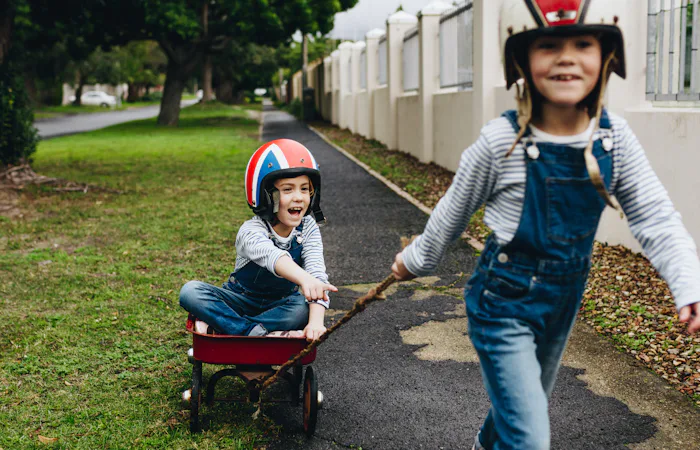 Little girl in a toy cart being pulled by her twin sister outside on road. Identical twins playing o...