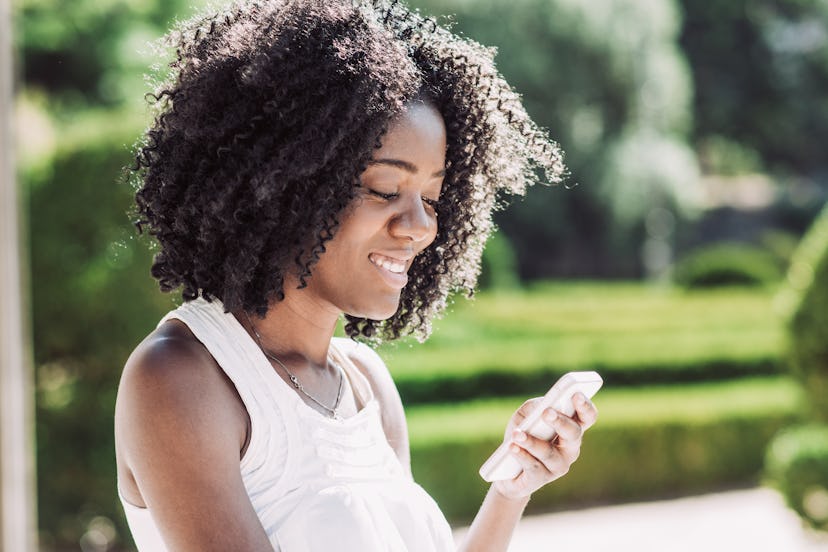 Smiling pretty young black woman texting on smartphone in park. Communication concept. Side closeup ...