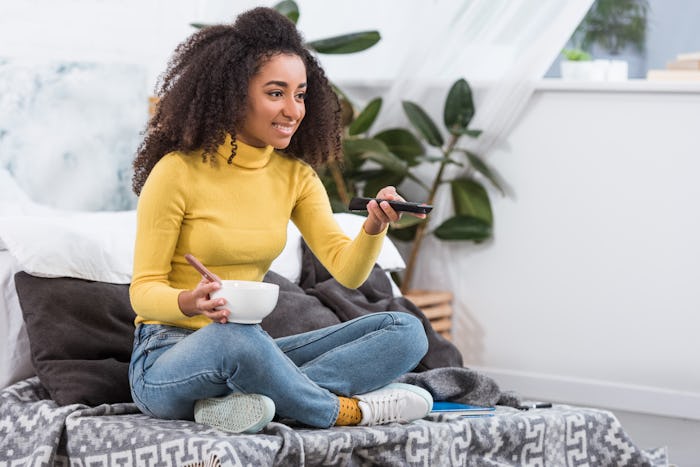 attractive stylish african american girl holding bowl with food and watching tv at home