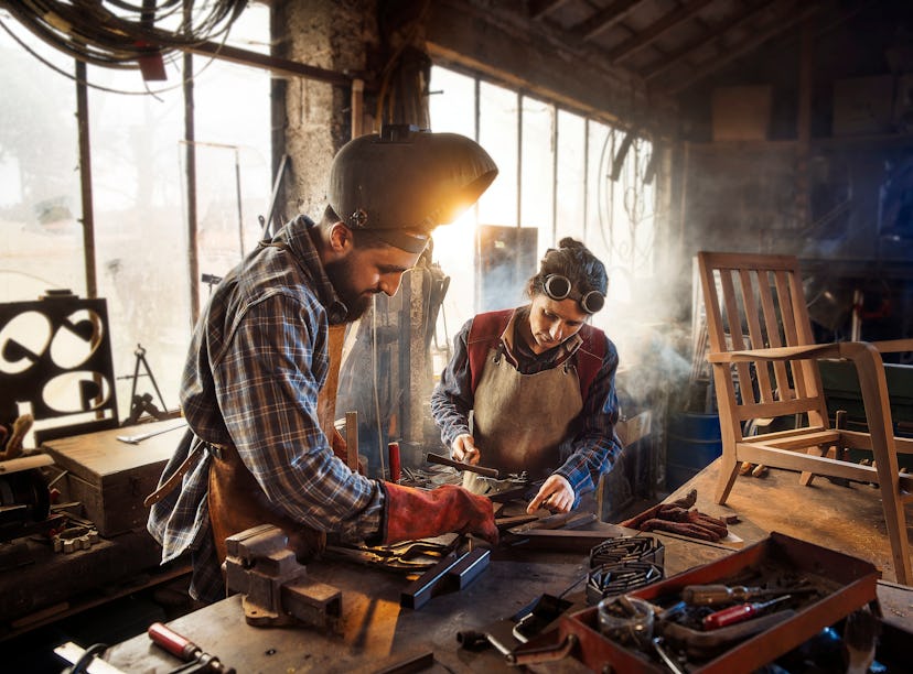 Two artisans welder in their craft workshop welding metal parts to assemble them on a designer woode...
