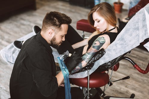 Professional tattooer doing tattoo on hand by tattoo machine while girl thoughtfully watching proces...