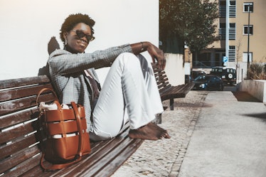 A young charming fancy African female in a plaid jacket and white trousers is sitting barefoot on a ...