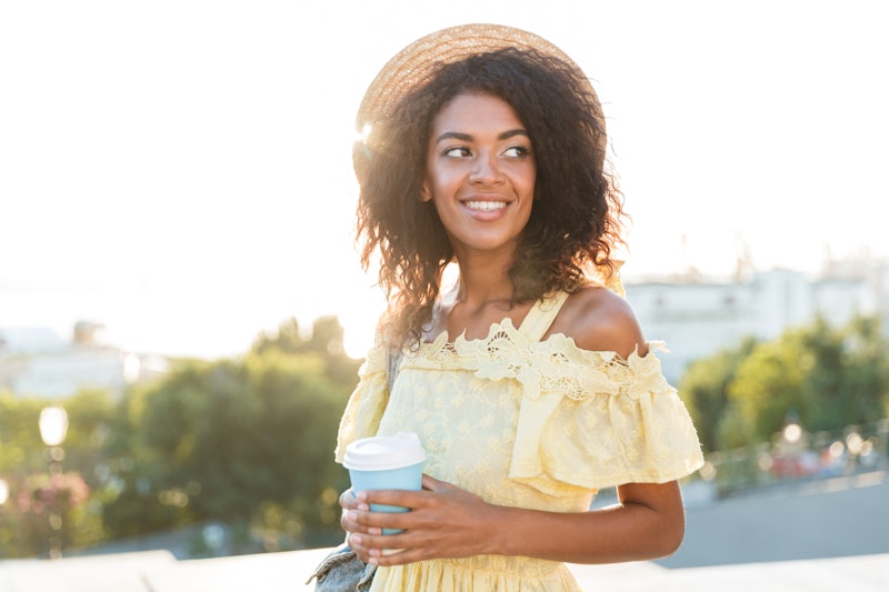 Happy young african woman in summer dress walking at the city, holding cup of coffee