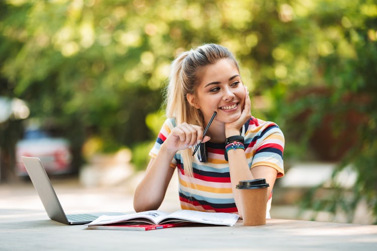 Image of a happy young cute girl student sitting in park using laptop computer writing notes.