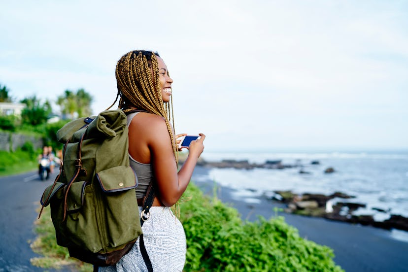Cheerful dark skinned female traveller holding smartphone device in hands while trekking around natu...
